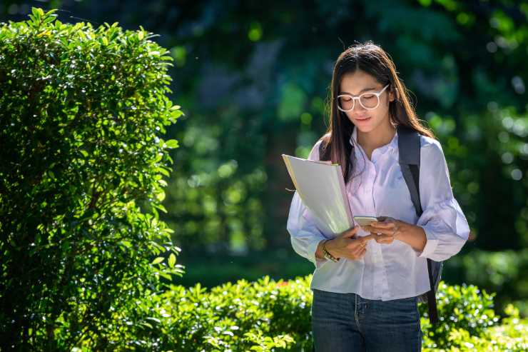 studentessa cammina con fogli in mano e cellulare circondata da piante