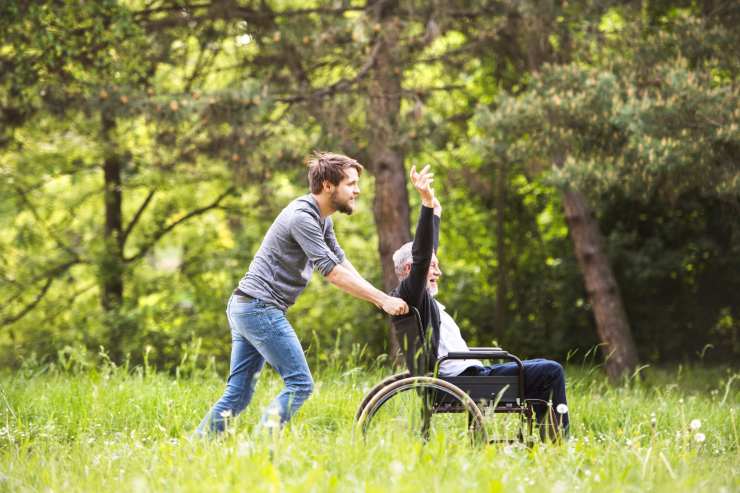 ragazzo spinge carrozzella disabile che alza felice le braccia al cielo